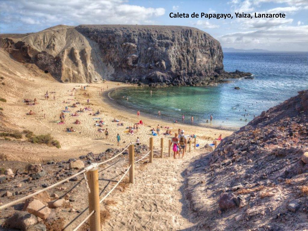 6-Caleta de Papagayo (Yaiza, Lanzarote) El sureste de Lanzarote se ve dorado en su arena, se siente frío por un Atlántico de tonos azules verdosos, sabe a salitre, suena a viento alisio y huele a arena quemada por el sol africano. Con Fuerteventura y el islote de Lobos al fondo, la caleta de Papagayo, la más fotogénica y aplacerada, la que da nombre al resto de playas del monumento natural de los Ajaches, sirvió hasta mediados del siglo pasado como fondeadero. Los niños gustan de su media luna bien trazada; otros practican snorkel en sus laterales. Pero todo se revela secundario frente a la panorámica que se divisa desde la terraza natural que otorga el acantilado, junto a El Chiringuito. El placer visual encuentra durante el crepúsculo su máxima expresión.